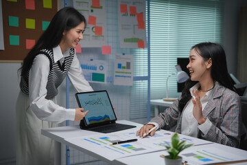 Two smiling businesswomen are discussing work while analyzing financial charts on a tablet in a bright office