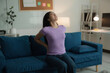 © amnaj - Young woman is sitting on her sofa, holding her back in pain, with her home office in the background