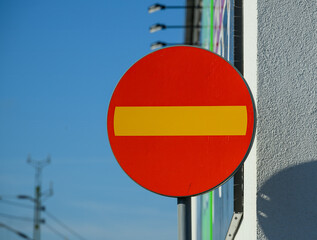 A bright red no entry sign with a horizontal yellow bar is mounted on a building. The clear blue sky serves as a backdrop, enhancing visibility during daylight hours