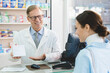 © InsideCreativeHouse - White blank medicine box container with drugs, vitamins, pills, painkillers, antibiotics. Middle-aged mature male pharmacist showing advising medications to female client customer