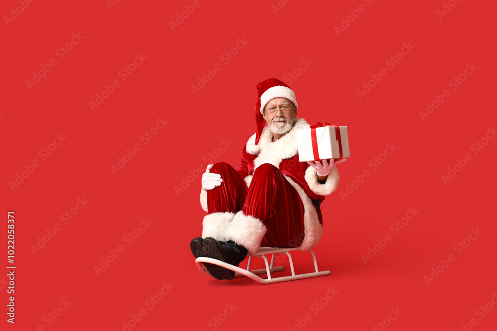Santa Claus in glasses with gift box sitting on sledge against red background