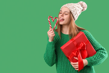 Happy young woman with candy canes and Christmas gift on green background