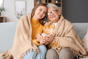 Wall Mural - Happy senior woman and her granddaughter with plaid drinking tea on sofa at home