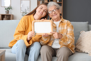 Wall Mural - Happy senior woman and her granddaughter with blank photo frame sitting on sofa at home