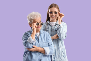 Wall Mural - Happy senior woman and her granddaughter in sunglasses on purple background