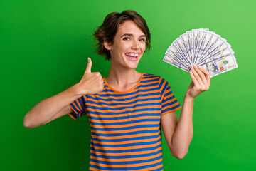 Wall Mural - Photo portrait of pretty young girl hold banknotes fan thumb up dressed stylish striped outfit isolated on green color background