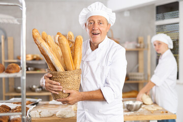 During work, elderly male baker in white uniform and apron demonstrates finished products, basket of baguettes. Business idea, production of bread products according to authors recipe