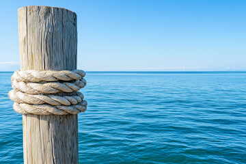 Close-up of a thick rope tied around a wooden dock post with a calm, blue ocean and clear sky in the background