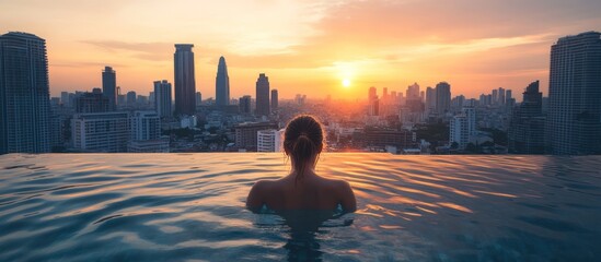 Wall Mural - Woman in infinity pool with city skyline and sunset in the background.