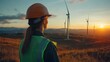 © Helois - Female engineer in hardhat and safety vest looks at wind turbines during sunset.