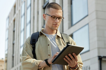 Poster - Medium portrait of man with glasses engaged in using tablet computer looking at screen while commuting to workplace or working outside in sunlight, copy space