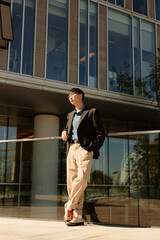 Vertical shot of Asian male corporate worker wearing stylish outfit for office leaning on glass construction posing in laid back way while relaxing in sunlight outside business building, copy space