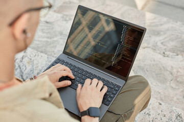 Over shoulder shot of unrecognizable male IT programmer writing code on laptop typing on computer keyboard while sitting on concrete steps outside in city street, copy space