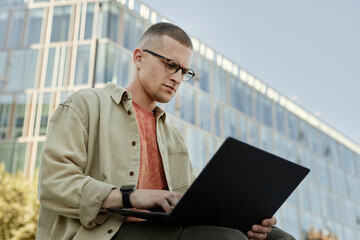 Poster - Medium shot of serious male IT programmer with glasses and tiny septum using laptop working outdoors while sitting outside in city street, copy space