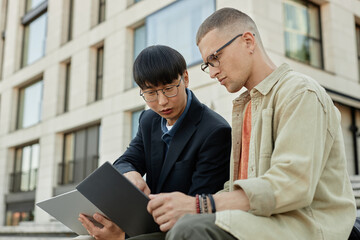 Medium shot of classy male Asian software developer with glasses assisting junior IT specialist pointing at laptop screen while collaborating outside in city, copy space