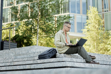 Poster - Full shot of concentrated male IT programmer in casual clothing using laptop to study or write code sitting on concrete steps with backpack lying next to man outside in city, copy space