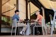 © Seventyfour - Side view of two adult male IT programmers using laptops to write code discussing work strategy while sitting at table in outdoor seating area of cafe, copy space