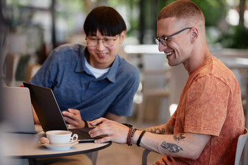 Side view of smiling male software engineer typing on laptop writing code while having fun working collaboratively with colleague during meeting over coffee in outdoor seating area of cafe, copy space