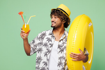 Young happy Indian man wear beach shirt hat rest near hotel pool hold rubber ring drink orange citrus cocktail look aside isolated on plain green color background. Summer vacation sea sun tan concept.