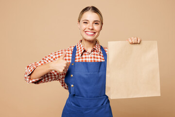 Wall Mural - Young woman barista bartender barman in blue apron shirt work in coffee shop hold clear blank craft paper takeaway bag show thumb up isolated on plain beige background Small business startup concept