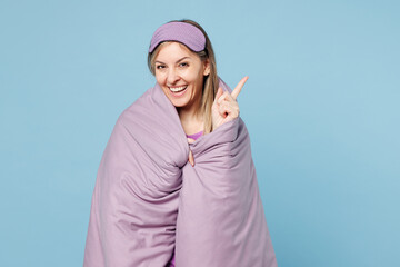 Wall Mural - Calm young woman wear purple pyjamas jam sleep eye mask wrapped in duvet rest relax at home point index finger aside on area isolated on plain pastel light blue background Good mood night nap concept