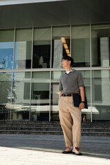 Poster - Vertical shot of fashionable Asian male IT programmer in office clothing holding laptop standing in sunlight next to business building while posing with hand in pocket outside in street, copy space