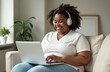 © Yliya - A large African American woman is sitting on the couch and working online on a laptop