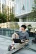 © Seventyfour - Vertical shot of Asian man looking away during work on laptop sitting on concrete sidewalk outside in city street while leaning against glass construction with building in background, copy space