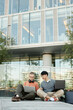 © Seventyfour - Vertical shot of diverse team of two male IT programmers working on laptops collaboratively sitting on pavement outside in city street against business building, copy space