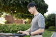 © Seventyfour - Medium shot of Asian male IT programmer in wired headphones concentrated on using laptop typing on keyboard while working at stand up desk in city park on blurred background, copy space