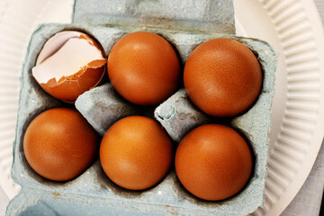 six golden brown chicken eggs in a pale blue egg box with one broken egg