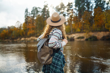 Female traveler with a backpack and a hat enjoying autumn hiking along the river. Active lifestyle. Hiking