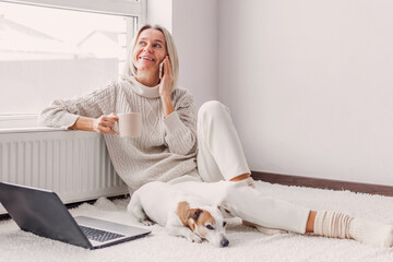 Wall Mural - Happy middle aged woman with dog using her laptop at cozy white home