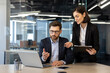 © Liubomir - Colleagues in formal attire engaged in business meeting. Woman pointing at laptop screen, man contemplating solution. Office desk with laptop, smartphone, and documents enhancing collaborative