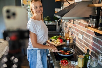 Wall Mural - Smiling young woman cooking and filming with smartphone in modern kitchen