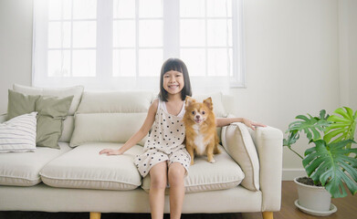 Portrait of little asian girl stay home sit on the sofa  living room with lovely dog. Happy moment of pure love toddler playing with little dog, minimal happy pet animal lifestyle concept