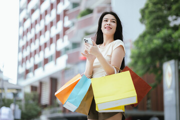 A cheerful young Asian woman, stylishly dressed, smiles excitedly while holding shopping bags. Enjoying her summer shopping experience, she embraces the fun and privilege of retail therapy, surrounded