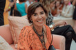 © Sascha - Photo of an elegant brunette Brazilian woman in her late thirties, wearing a coral dress. She has short brown hair, sitting on the couch at fashion week after a show