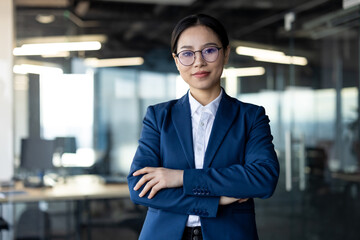 Asian business woman confidently standing in office, arms crossed, wearing glasses and suit. Emphasizes professionalism, leadership, corporate success, female empowerment in modern workplace.