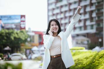 Active Asian woman in suit standing outdoors in city holding smartphone and laptop. She represents the future of finance and investment, keeping updated on news and opportunities.