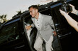 © Seventyfour - Dutch angle shot of smiling well off man wearing grey formal attire caught by paparazzi getting out of limousine at award ceremony arrival, camera flash, copy space