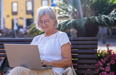 Wall Mural - Caucasian senior woman sitting outdoor on a bench using laptop. Grey haired elderly lady relax enjoying tech and social