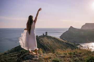 Wall Mural - A woman in a white dress stands on a rocky hill overlooking the ocean. She is smiling and she is happy.