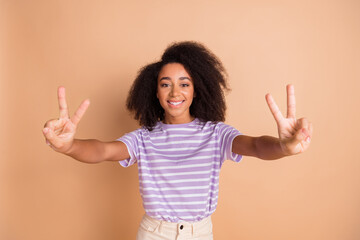 Poster - Portrait of pretty young girl demonstrate v-sign wear striped t-shirt isolated on beige color background