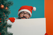 © dargog - Man dressed in a festive Santa Claus costume in glasses holding blank with christmas tree background copy space and mockup.