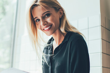 Wall Mural - Portrait of cheerful female student with perfect white teeth smiling at camera during time for planning course work parts, happy hipster girl in casual wear holding textbook for education indoors