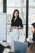 © BullRun - Portrait of confident business woman standing near flip chart for presentation and looking at camera, young male and female colleagues using white board with copy space area for your advertising