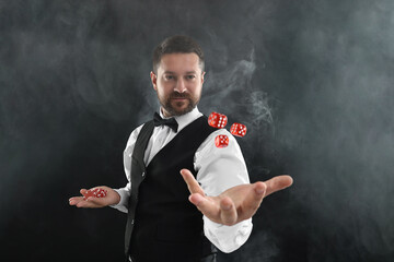 Wall Mural - Professional croupier holding dice and casino chips on black background with smoke