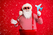 © deagreez - Photo of old man grey beard demonstrate blue paper plane funny wear santa claus x-mas costume suspenders sunglass striped shirt cap isolated red color background