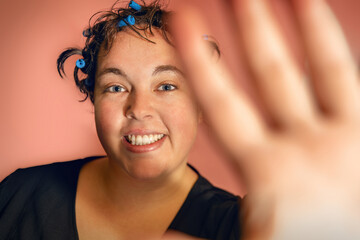 A young woman doing her hair with hair curler in front of colorful pink studio background; body positivity concept